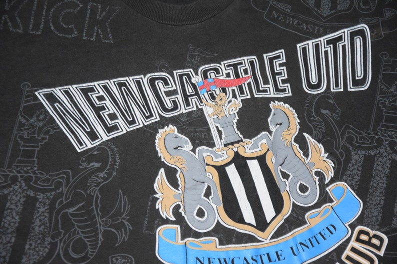K&ouml;nnte beinhalten: Schwarzes T-Shirt mit einem wei&szlig;-schwarzen Logo des Newcastle United Football Clubs. Das Logo zeigt ein Wappen mit zwei Seepferden, einem Schloss und einem Banner mit der Aufschrift "Newcastle United".