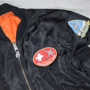 May include: Black bomber jacket with a zipper closure and two embroidered patches. One patch features a red circle with a white cross and the text "Swiss Hornet Team". The other patch features a blue and white airplane with the text "Patrouille Suisse".