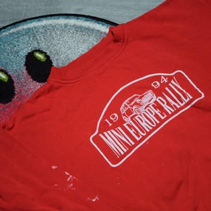 Może przedstawiać: Czerwona bluza z białym nadrukiem z napisem "1994 MINI EUROPE RALLY" z ilustracją samochodu. Bluza jest lekko pomarszczona i ma białe ślady. Tło obejmuje kolorowy dywan.