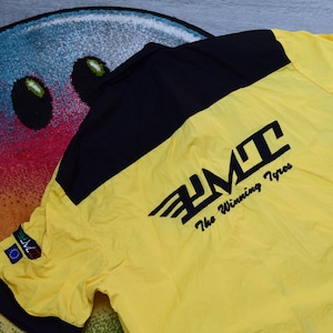 Pode incluir: Casaco amarelo e preto de manga curta com o texto "PMT The Winning Tyres". O casaco está colocado sobre um tapete colorido com um desenho de rosto sorridente. O casaco tem gola preta e detalhes pretos nos ombros.