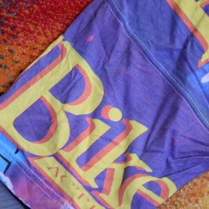 Puede incluir: Una camiseta morada, rosa, azul y amarilla con la palabra "Bike" impresa en amarillo con un contorno rosa. La palabra "Active" est&aacute; impresa en letras m&aacute;s peque&ntilde;as debajo de la palabra "Bike".