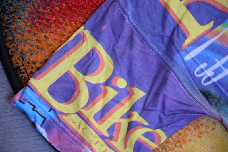 Puede incluir: Una camiseta morada, rosa, azul y amarilla con la palabra "Bike" impresa en amarillo con un contorno rosa. La palabra "Active" est&aacute; impresa en letras m&aacute;s peque&ntilde;as debajo de la palabra "Bike".