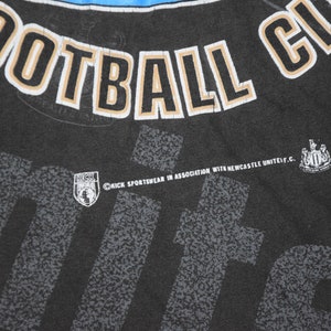 K&ouml;nnte beinhalten: Schwarzes T-Shirt mit einem wei&szlig;en und goldenen "Newcastle United Football Club"-Motiv. Das Shirt hat au&szlig;erdem ein kleines Logo f&uuml;r "Kick Sportswear in association with Newcastle United F.C."