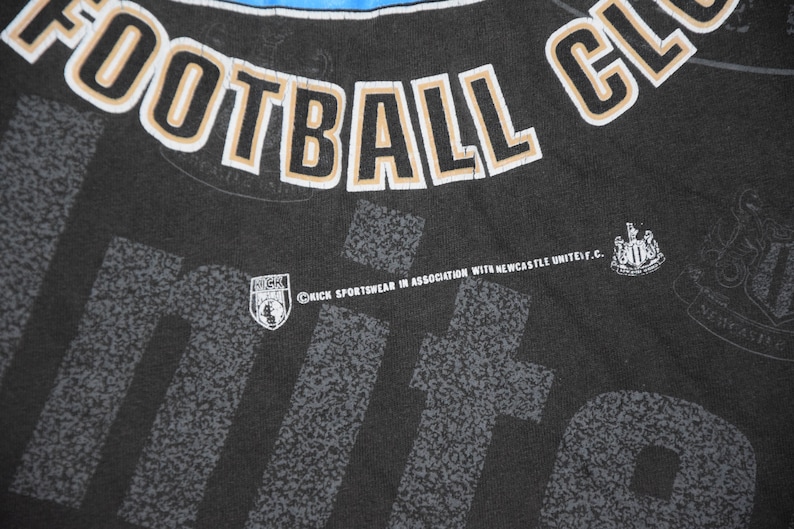 K&ouml;nnte beinhalten: Schwarzes T-Shirt mit einem wei&szlig;en und goldenen "Newcastle United Football Club"-Motiv. Das Shirt hat au&szlig;erdem ein kleines Logo f&uuml;r "Kick Sportswear in association with Newcastle United F.C."