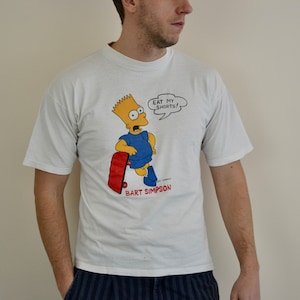 May include: White t-shirt with a cartoon image of Bart Simpson skateboarding and a speech bubble that says "Eat my shorts!" The image is in color and includes the text "Bart Simpson".