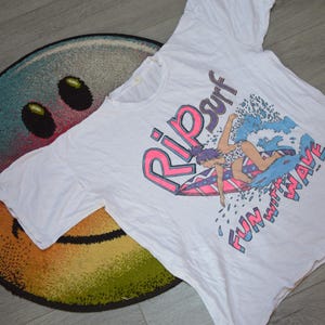 Op de afbeelding: Wit T-shirt met een kleurrijke afbeelding van een surfer op een golf, met de woorden "Rip Surf" en "Fun with Wave". Het T-shirt ligt op een ronde, veelkleurige smiley-vloerkleed.