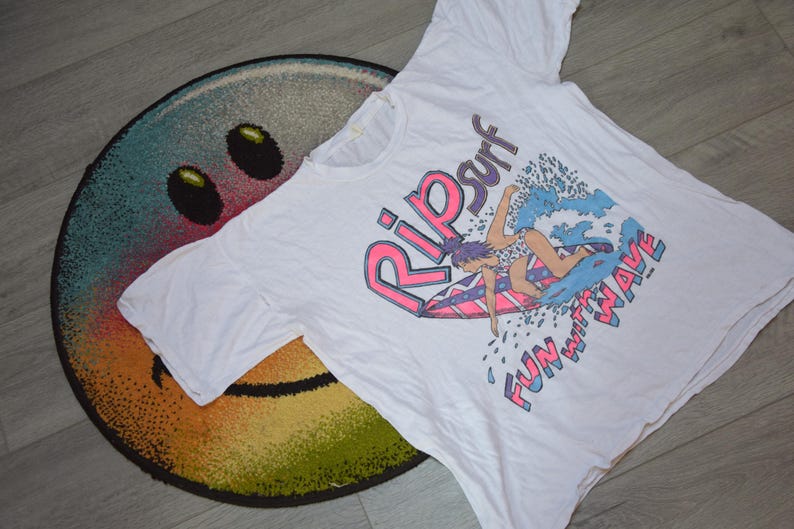 Op de afbeelding: Wit T-shirt met een kleurrijke afbeelding van een surfer op een golf, met de woorden "Rip Surf" en "Fun with Wave". Het T-shirt ligt op een ronde, veelkleurige smiley-vloerkleed.
