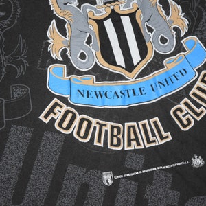 K&ouml;nnte beinhalten: Schwarzes T-Shirt mit einem wei&szlig;-schwarzen Logo des Newcastle United Football Club. Das Logo zeigt einen Schild mit drei vertikalen Streifen, zwei Seepferdchen und einem Banner mit der Aufschrift "Newcastle United".