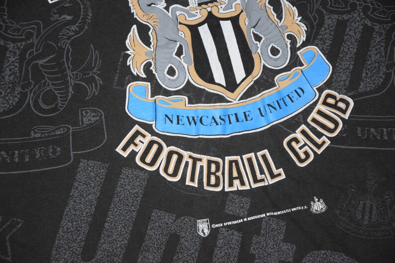 K&ouml;nnte beinhalten: Schwarzes T-Shirt mit einem wei&szlig;-schwarzen Logo des Newcastle United Football Club. Das Logo zeigt einen Schild mit drei vertikalen Streifen, zwei Seepferdchen und einem Banner mit der Aufschrift "Newcastle United".