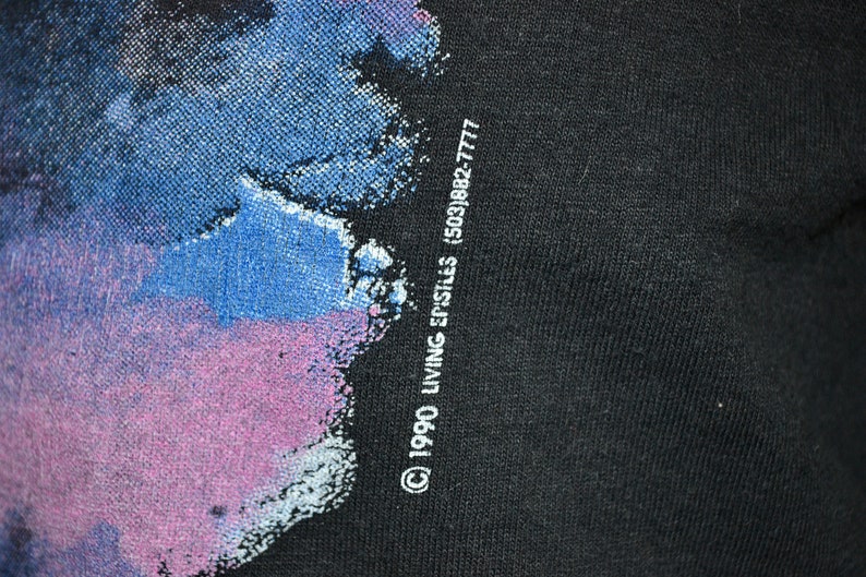 Może przedstawiać: Czarny t-shirt z kolorowym abstrakcyjnym wzorem w odcieniach niebieskiego, fioletowego i białego. Tekst "&copy; 1990 LIVING EPISTLES (503)882-7777" jest wydrukowany na dole po prawej stronie.