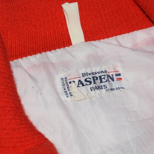 May include: A white and red jacket with a white label that says "Blousons Aspen Paris (1) 880-3323" in black text.