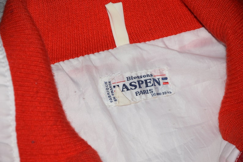 May include: A white and red jacket with a white label that says "Blousons Aspen Paris (1) 880-3323" in black text.