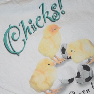 May include: White t-shirt with a teal graphic that says "Chicks! Born to Play". The graphic also features three yellow chicks with soccer balls.
