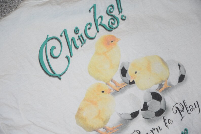 May include: White t-shirt with a teal graphic that says "Chicks! Born to Play". The graphic also features three yellow chicks with soccer balls.