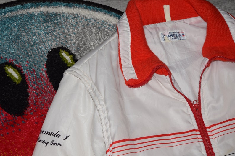 May include: White windbreaker jacket with red stripes and a red collar. The jacket has a zipper closure and the text "Formula 1 Racing Team" on the left sleeve. The label on the jacket reads "Aspen Paris".