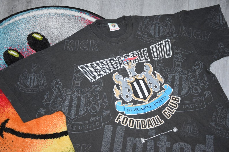 K&ouml;nnte beinhalten: Schwarzes T-Shirt mit einem wei&szlig;en Allover-Druck des Logos des Newcastle United Football Clubs und den Worten "Newcastle Utd" und "Football Club".