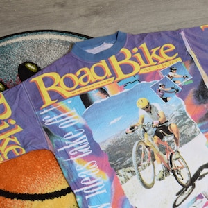Puede incluir: Una camiseta morada, azul y amarilla con un estampado gr&aacute;fico de una persona montando una bicicleta de monta&ntilde;a. El texto "Road Bike Action" est&aacute; impreso en la camiseta.