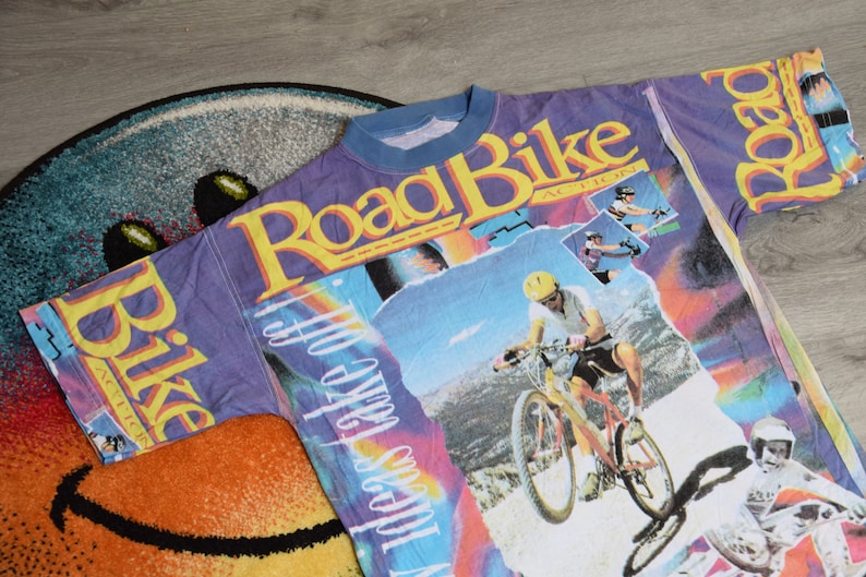 Puede incluir: Una camiseta morada, azul y amarilla con un estampado gr&aacute;fico de una persona montando una bicicleta de monta&ntilde;a. El texto "Road Bike Action" est&aacute; impreso en la camiseta.