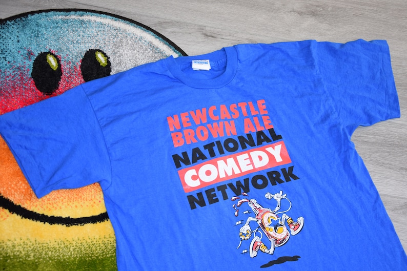 K&ouml;nnte beinhalten: Ein blaues T-Shirt mit dem Text "NEWCASTLE BROWN ALE NATIONAL COMEDY NETWORK" in Rot, Schwarz und Wei&szlig;. Unter dem Text befindet sich eine Comicfigur mit einem Mikrofon. Das Shirt besteht aus einem weichen Material.
