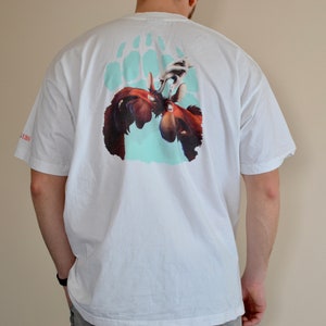 May include: White t-shirt with a graphic print of two moose facing each other inside a bear paw print. The moose are brown and have antlers. The bear paw print is light blue.