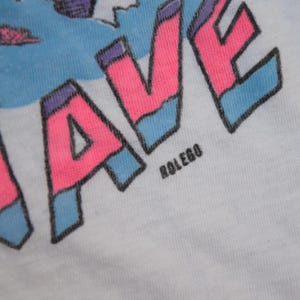 Op de afbeelding: Close-up van een wit t-shirt met een kleurrijke afbeelding. Het woord "WAVE" staat prominent in roze, omlijnd in zwart en geaccentueerd met blauw. De tekst "ROLEGO" is onder het woord "WAVE" gedrukt.