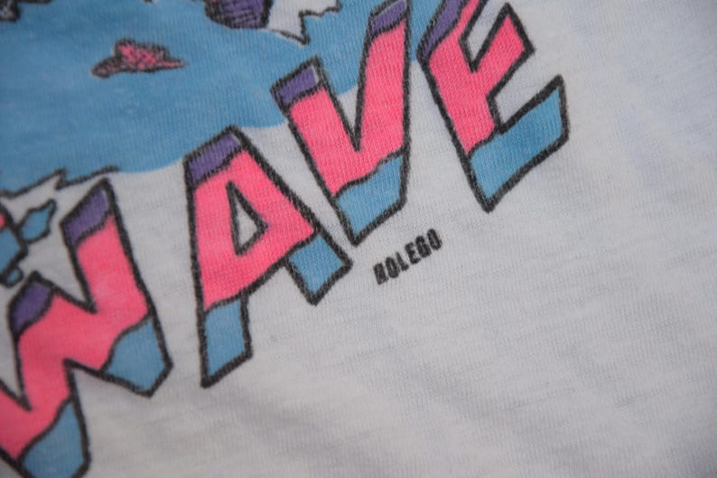 Op de afbeelding: Close-up van een wit t-shirt met een kleurrijke afbeelding. Het woord "WAVE" staat prominent in roze, omlijnd in zwart en geaccentueerd met blauw. De tekst "ROLEGO" is onder het woord "WAVE" gedrukt.