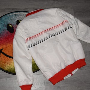 May include: White bomber jacket with red and black striped detail on the back. The jacket has a red ribbed collar and cuffs.
