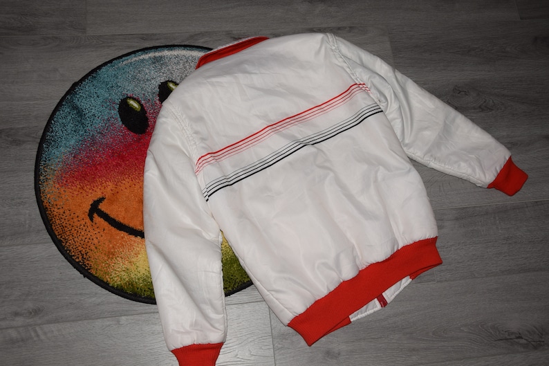May include: White bomber jacket with red and black striped detail on the back. The jacket has a red ribbed collar and cuffs.