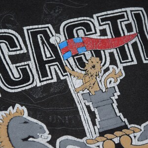 K&ouml;nnte beinhalten: Schwarzes T-Shirt mit einer wei&szlig;en Grafik eines L&ouml;wen, der eine Flagge mit roten, wei&szlig;en und blauen Streifen h&auml;lt. Der Text "CASTLE" ist in Wei&szlig; auf das Shirt gedruckt.