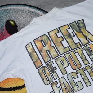 Puede incluir: Camiseta blanca con un estampado gráfico de las palabras "I REEK OF PUTRE FACTION" en negro y amarillo. El gráfico está formado por imágenes de personas.