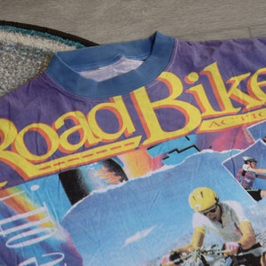 Puede incluir: Una camiseta morada, azul y amarilla con un estampado gr&aacute;fico de las palabras "Road Bike Action" e im&aacute;genes de personas montando en bicicleta.
