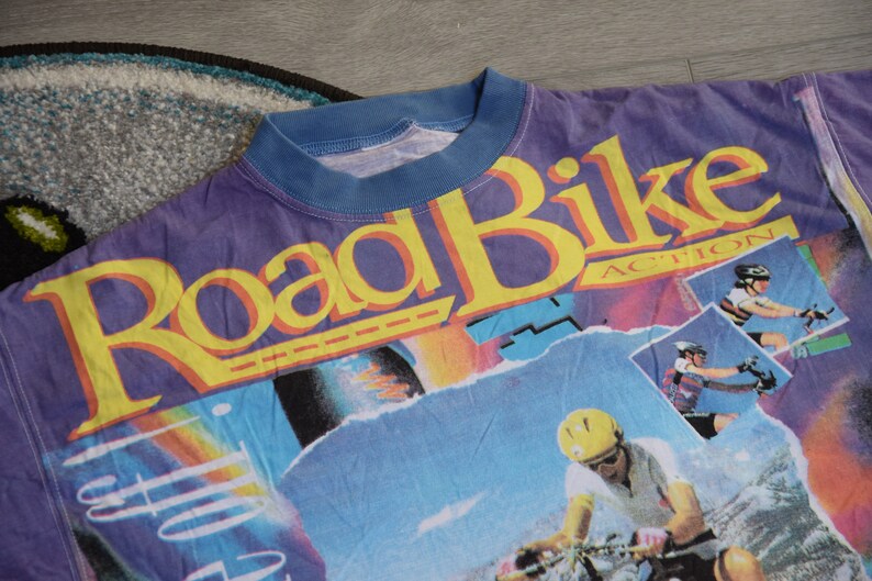 Puede incluir: Una camiseta morada, azul y amarilla con un estampado gr&aacute;fico de las palabras "Road Bike Action" e im&aacute;genes de personas montando en bicicleta.