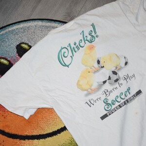 May include: White t-shirt with a graphic of baby chicks playing soccer. The text reads "Chicks! We're Born to Play Soccer. Power of a Girl."