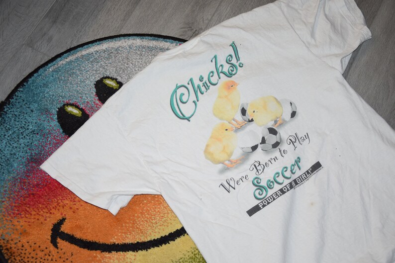 May include: White t-shirt with a graphic of baby chicks playing soccer. The text reads "Chicks! We're Born to Play Soccer. Power of a Girl."