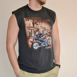 May include: Dark gray sleeveless t-shirt featuring a Harley-Davidson graphic. The graphic depicts a person riding a motorcycle in front of a building. The shirt has a crew neck and is made of a soft material.