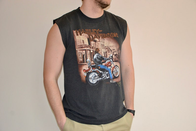 May include: Dark gray sleeveless t-shirt featuring a Harley-Davidson graphic. The graphic depicts a person riding a motorcycle in front of a building. The shirt has a crew neck and is made of a soft material.