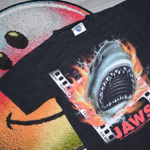 Puede incluir: Camiseta negra con un gráfico de un tiburón con llamas detrás. La camiseta tiene el texto "JAWS" en rojo y "AT UNIVERSAL STUDIOS FLORIDA" en blanco.