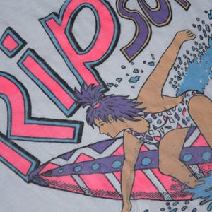 Op de afbeelding: Wit T-shirt met een neonroze en blauwe afbeelding van een surfer op een golf. Het woord "RIP" staat in grote roze letters met blauwe contouren. De surfer draagt een badpak met patroon.
