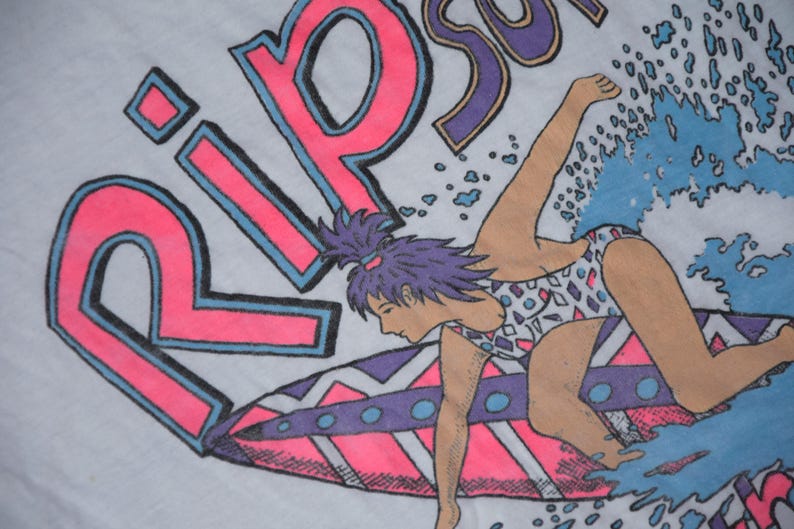 Op de afbeelding: Wit T-shirt met een neonroze en blauwe afbeelding van een surfer op een golf. Het woord "RIP" staat in grote roze letters met blauwe contouren. De surfer draagt een badpak met patroon.