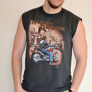 May include: Dark gray sleeveless t-shirt featuring a Harley-Davidson graphic. The graphic depicts a person riding a red motorcycle in front of a building. The Harley-Davidson logo is printed at the top of the graphic.