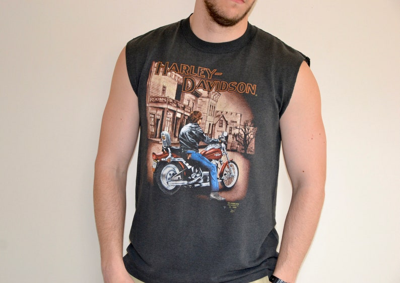 May include: Dark gray sleeveless t-shirt featuring a Harley-Davidson graphic. The graphic depicts a person riding a red motorcycle in front of a building. The Harley-Davidson logo is printed at the top of the graphic.