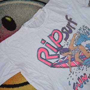 Op de afbeelding: Wit T-shirt met een surfafbeelding. Het ontwerp bevat een surfmeisje op een surfboard, de woorden "Rip Surf" in roze en paars, en de zin "Fun with Wave" in roze.
