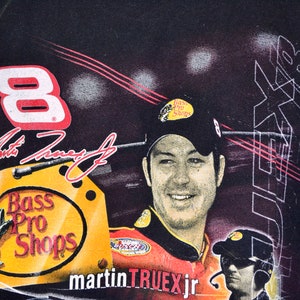Op de afbeelding: Zwart T-shirt met een portret van Martin Truex Jr. met een Bass Pro Shops pet. Het shirt toont nummer 8, de handtekening van Truex en het Bass Pro Shops logo. De achtergrond heeft een racethema.