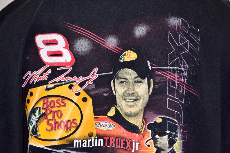 Op de afbeelding: Zwart T-shirt met een portret van Martin Truex Jr. met een Bass Pro Shops pet. Het shirt toont nummer 8, de handtekening van Truex en het Bass Pro Shops logo. De achtergrond heeft een racethema.