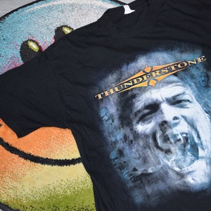 May include: Black t-shirt featuring a distressed graphic of a screaming face with the word "THUNDERSTONE" in orange and white. The shirt is made of a soft material and is likely a band or music-related item.