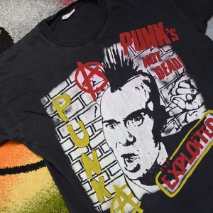 Pode incluir: Camiseta preta com uma estampa gráfica de um punk rocker com o texto "Punk's Not Dead!" e "The Exploited" em amarelo e vermelho.
