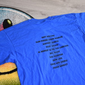 K&ouml;nnte beinhalten: Ein blaues T-Shirt mit schwarzem Text, der Namen wie Matt Welcome und Alan Parker-Urban Warrior auflistet. Das Shirt liegt auf einem bunten Teppich mit Smiley-Gesicht.