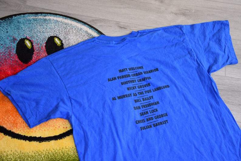 K&ouml;nnte beinhalten: Ein blaues T-Shirt mit schwarzem Text, der Namen wie Matt Welcome und Alan Parker-Urban Warrior auflistet. Das Shirt liegt auf einem bunten Teppich mit Smiley-Gesicht.