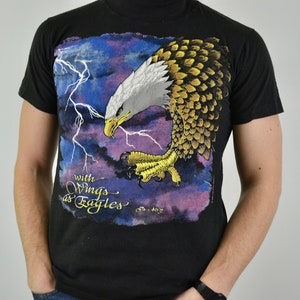 Może przedstawiać: Czarny t-shirt z szczeg&oacute;łową grafiką orła bielika z rozpostartymi skrzydłami, na tle błyskawic i fioletowo-niebieskiego nieba. Pod orłem znajduje się napis "with Wings as Eagles".