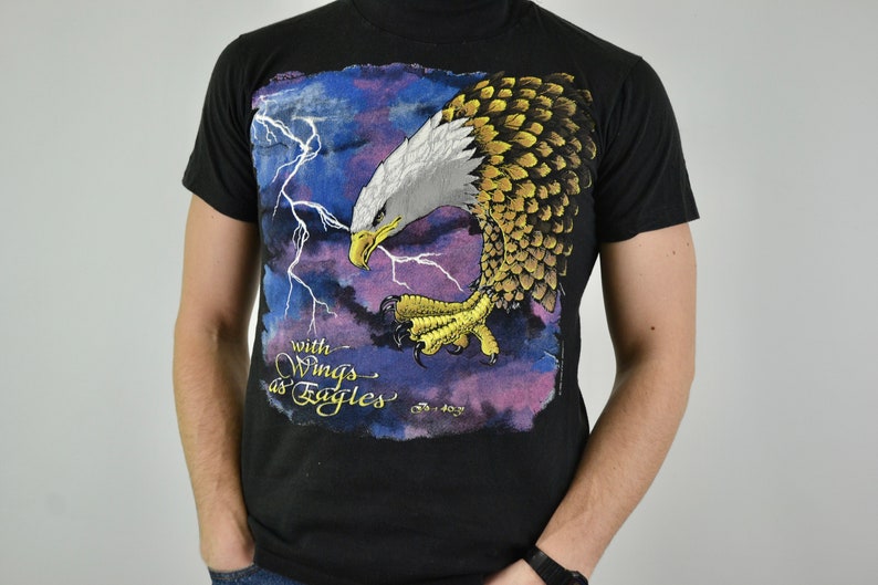 Może przedstawiać: Czarny t-shirt z szczeg&oacute;łową grafiką orła bielika z rozpostartymi skrzydłami, na tle błyskawic i fioletowo-niebieskiego nieba. Pod orłem znajduje się napis "with Wings as Eagles".